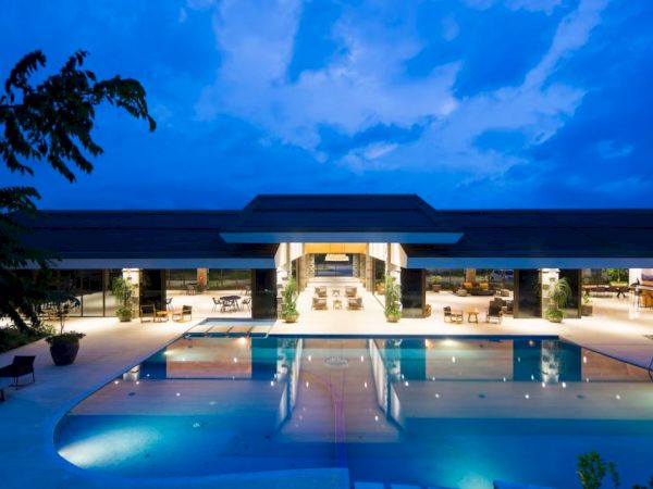 A luxurious building at night with a large illuminated pool, surrounded by seating areas and greenery under a deep blue sky.