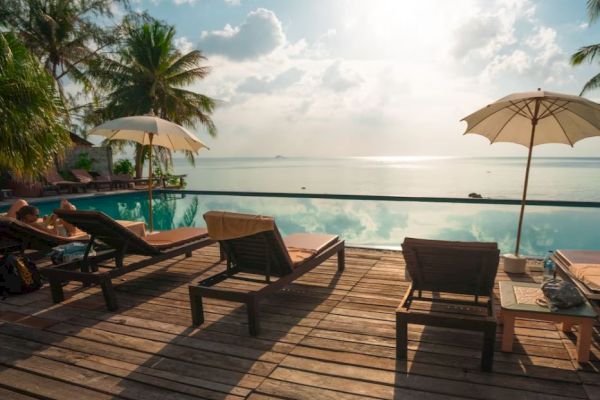 The image shows a relaxing poolside setup with lounge chairs, umbrellas, and a scenic ocean view under a partly cloudy sky.