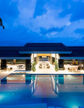 A luxurious modern building with large open spaces is reflected in a swimming pool, surrounded by lounge chairs, under a blue evening sky.