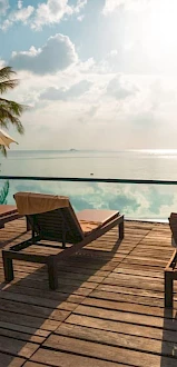 A serene poolside scene with lounge chairs, umbrellas, and palm trees overlooking a calm sea under a partly cloudy sky.