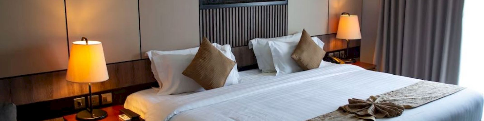 The image shows a neatly arranged hotel room with a large bed, decorative pillows, warm lighting, and modern decor.