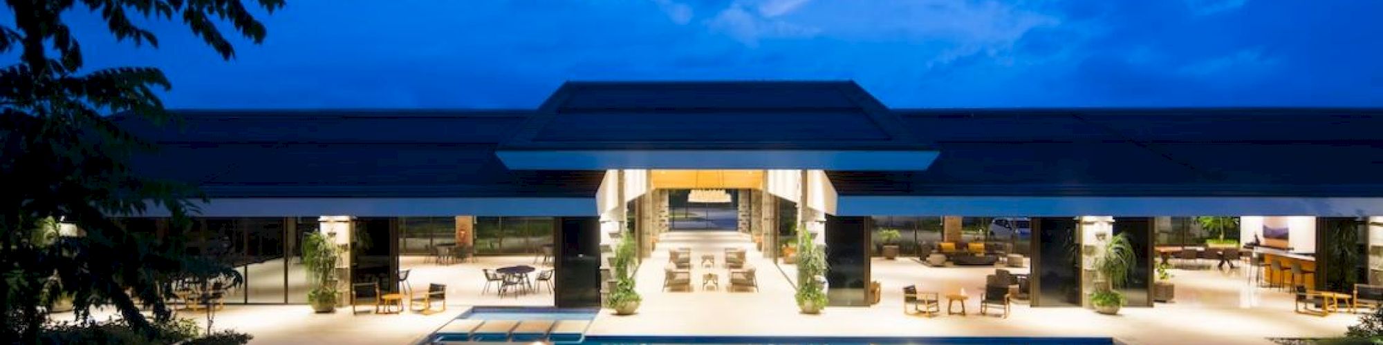 A luxurious building with open-air design, featuring a large swimming pool in the foreground under a twilight sky, surrounded by lounge chairs.