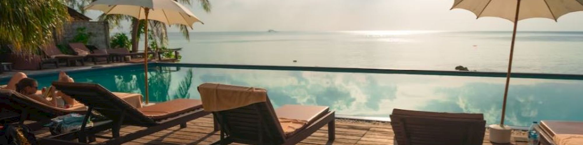 Wooden deck with lounge chairs and umbrellas by a pool, overlooking a tranquil ocean view with palm trees and sunlight.
