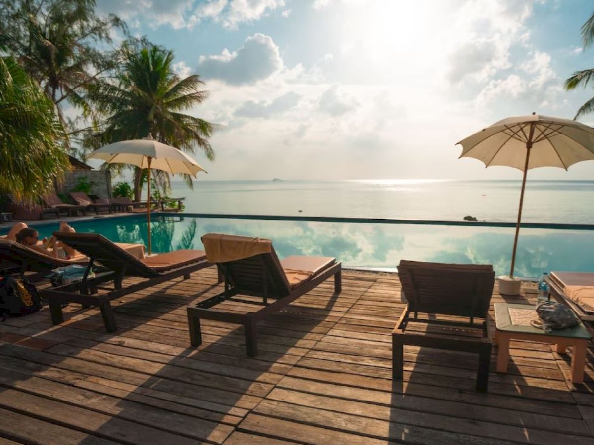 The image shows a serene poolside with lounge chairs, umbrellas, trees, and a view of the ocean under a partly cloudy sky.