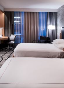 A modern hotel room with two beds, a flat-screen TV, desk, and chairs, featuring contemporary decor and lighting.