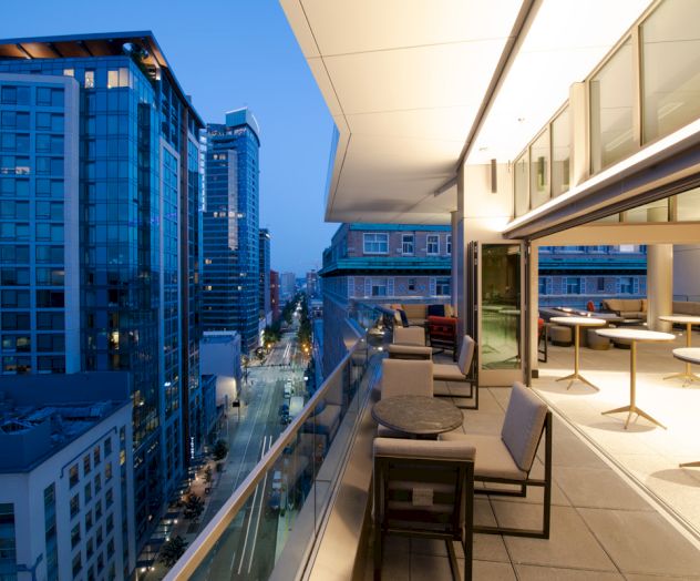 A modern cityscape at dusk with tall buildings and a spacious balcony featuring seating and tables, overlooking a lit-up urban street.
