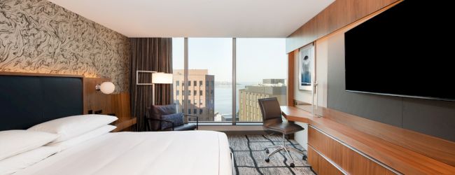 A modern hotel room with a large bed, a desk by the window, and a flat-screen TV on the wall, overlooking city buildings.