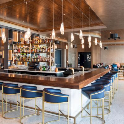 A modern bar with a central counter, blue chairs, pendant lights, and stocked shelves, surrounded by stylish seating and decor.