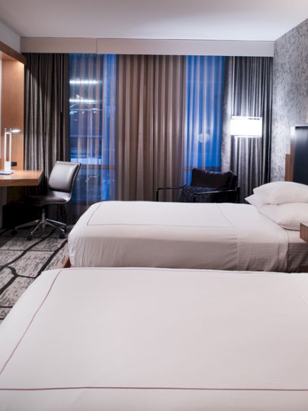 A modern hotel room with two beds, a desk, a TV, and stylish lighting. Curtains cover a large window.