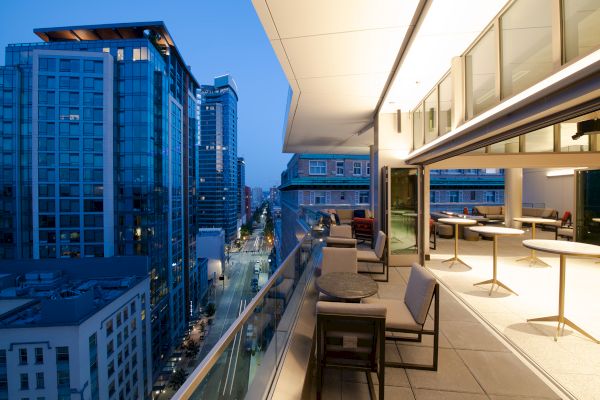 A modern rooftop terrace with seating, overlooking a cityscape of tall buildings and a street lit up at dusk.