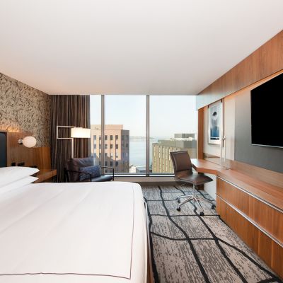 A modern hotel room with a large bed, wall-mounted TV, desk, and window with a city view. The decor is sleek and contemporary.