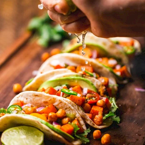 Tacos filled with chickpeas, diced vegetables, and garnished with avocado and cilantro, being sprinkled with lime juice.