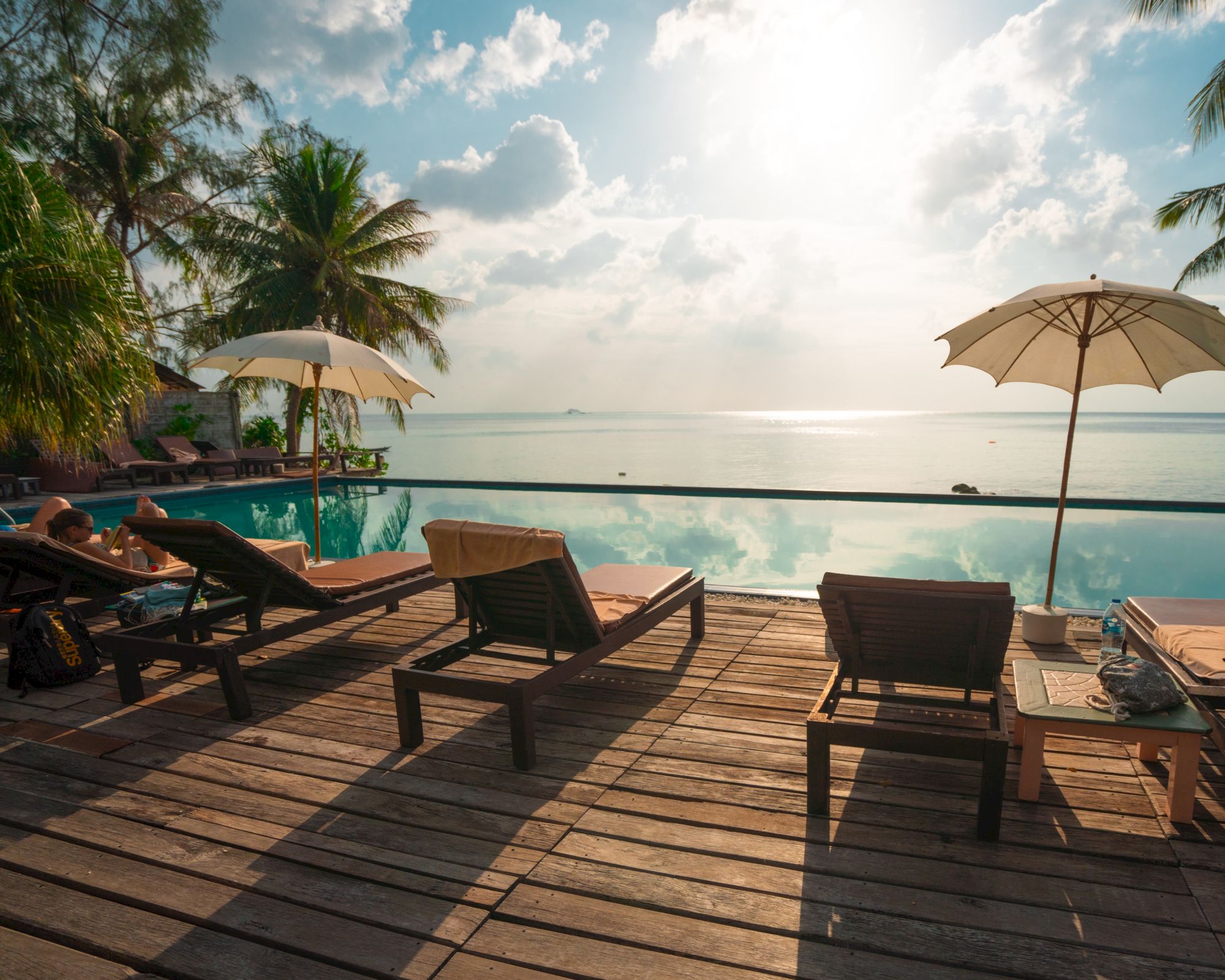 Lounge chairs and umbrellas on a wooden deck overlook a tranquil infinity pool and ocean, with lush palm trees and a bright sky in the background.