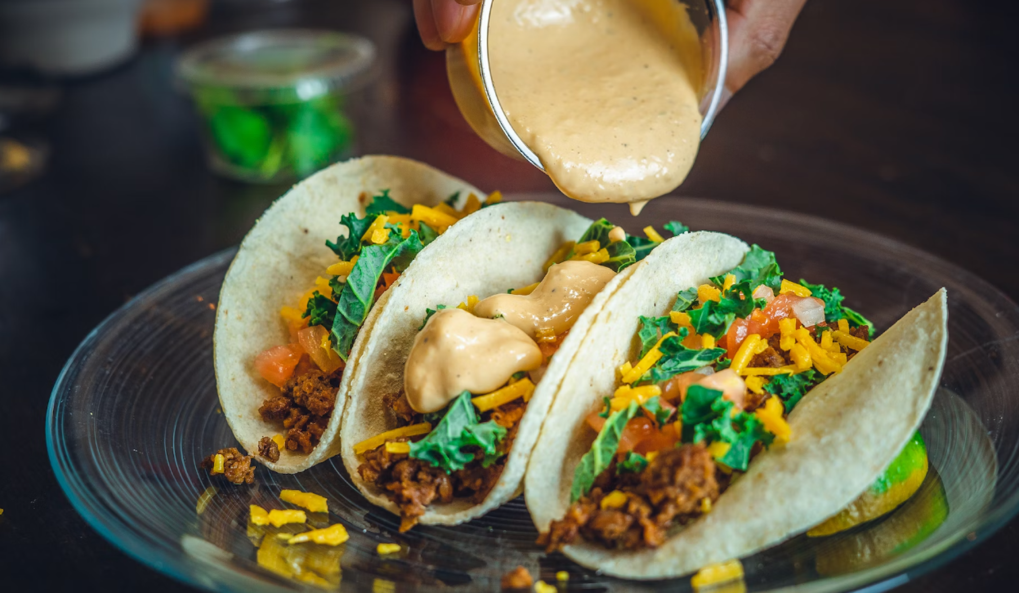 Three tacos filled with greens, cheese, and tomato, being topped with a creamy sauce on a clear plate.