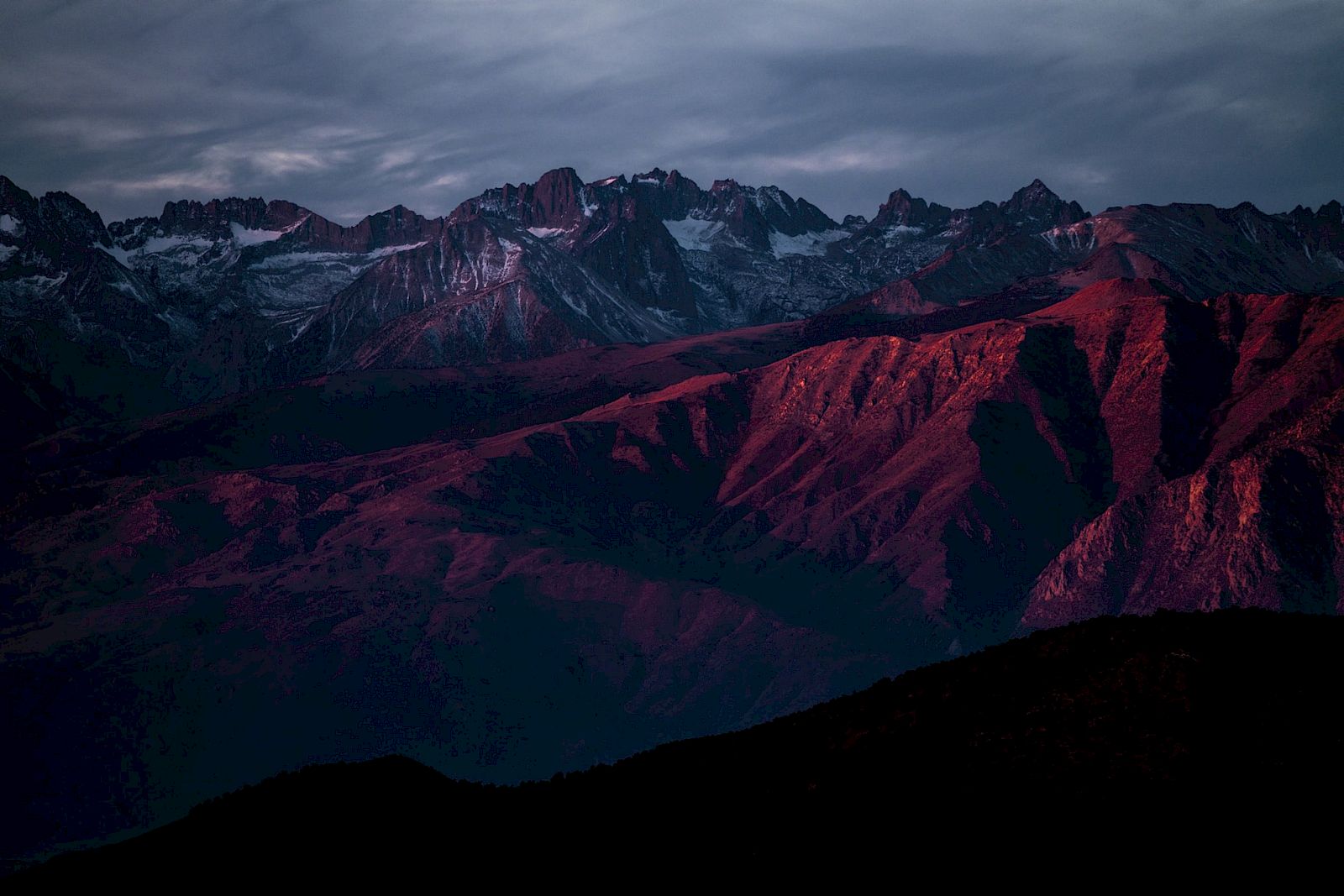 A mountain range with snowy peaks under a cloudy sky, bathed in a red sunset glow.