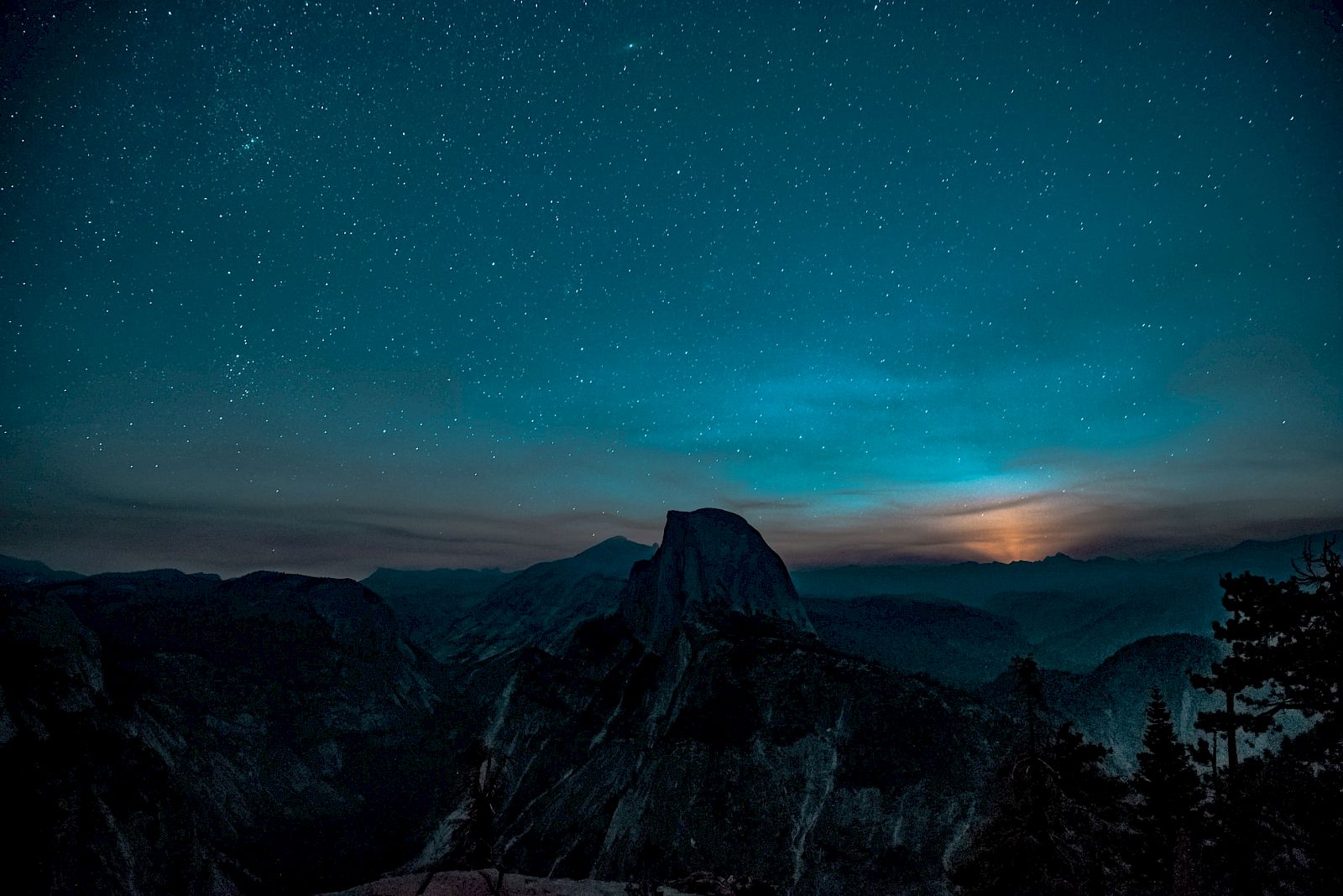 A starry night sky over a mountainous landscape with a faint glow on the horizon and silhouettes of trees in the foreground.