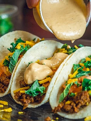 Three tacos filled with ground meat, cheese, lettuce, and tomatoes on a plate. Someone is pouring sauce over the tacos.