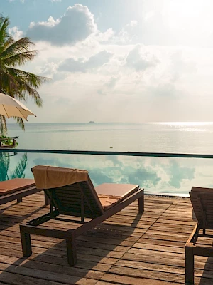 Poolside view with deck chairs and umbrellas facing a calm ocean, surrounded by palm trees, under a partly cloudy sky.