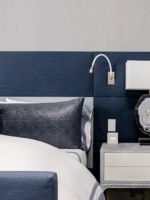 A modern bedroom features a blue headboard, white bedding, two pillows, a nightstand with a lamp, and a wall-mounted reading light.