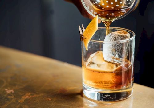 A drink is being poured into a glass with ice and garnished with a citrus peel on a table.