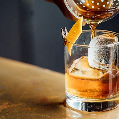 A drink is being poured into a glass with ice and garnished with a citrus peel on a table.