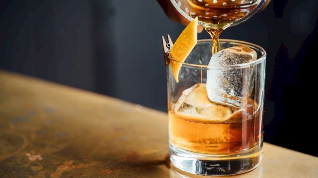 A drink is being poured into a glass with ice and garnished with a citrus peel on a table.