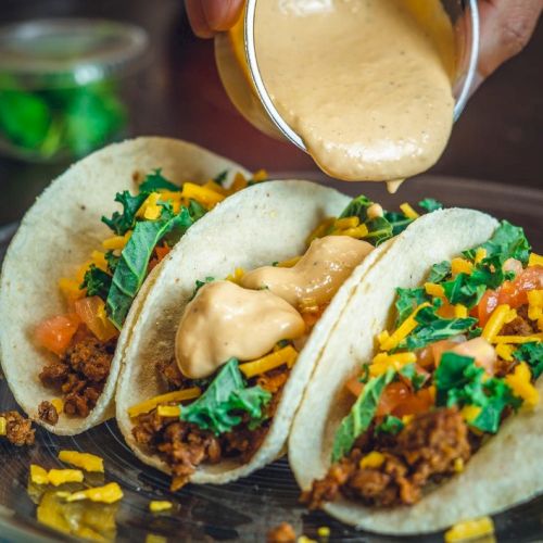 Three tacos with meat, cheese, lettuce, and tomatoes are being topped with a creamy sauce on a plate.