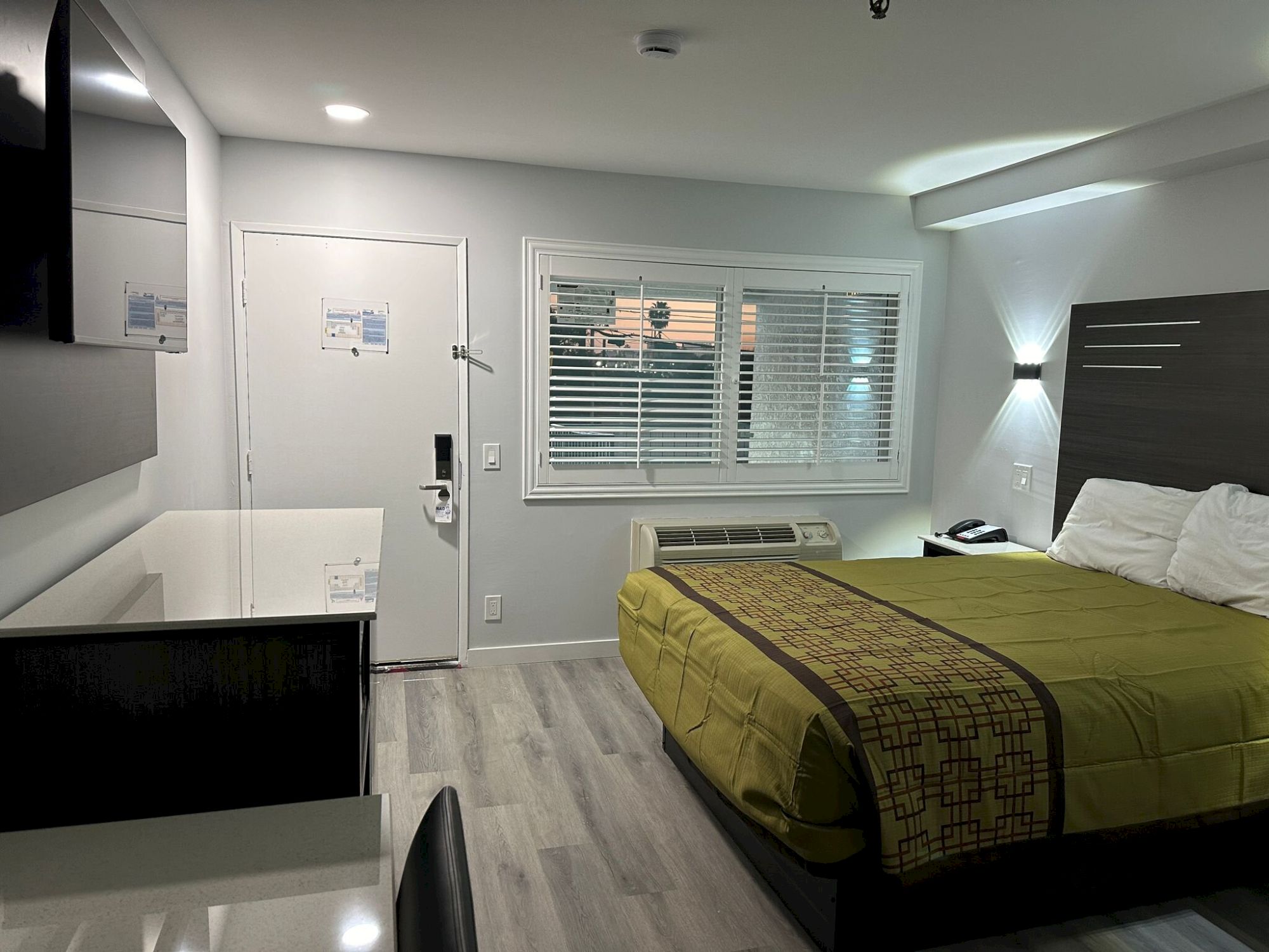 A modern, tidy hotel room with a bed, TV, desk, and window. White walls and wooden floors create a minimalist vibe.