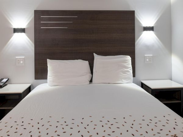 A neatly made bed with a wooden headboard, wall-mounted lights, and a white bedspread with a pattern. Two bedside tables complete the setup.