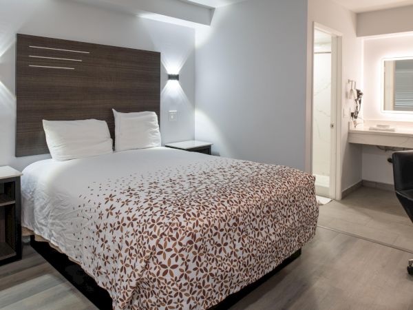A modern hotel room with a double bed, patterned duvet, side tables, and a bathroom vanity area. Simple and well-lit ambiance.
