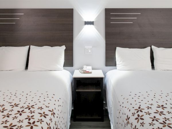 The image shows two neatly made beds with patterned sheets, a central nightstand, and a wall light above in a hotel room setting.