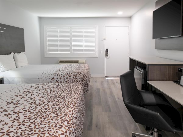 The image shows a modern hotel room with two beds, a desk, a wall-mounted TV, and a window with blinds.