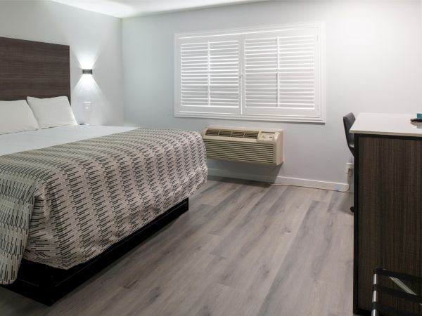 A modern hotel room with a bed, white walls, wooden floor, window with blinds, air conditioner, desk, and TV.