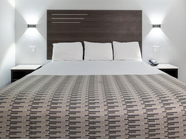 This image shows a neatly made bed with a geometric patterned bedspread, three pillows, modern wall lamps, and dark wooden headboard.