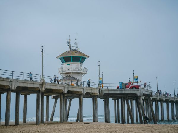 A coastal pier extends over the ocean with a lifeguard tower in view, surrounded by lampposts and some people walking along.