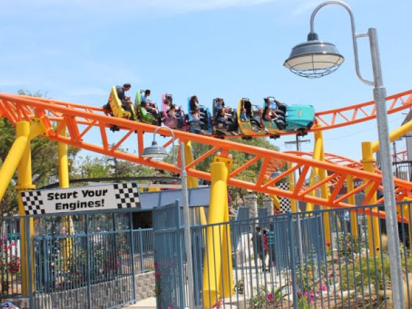 A roller coaster with riders is seen on an orange track above a fenced area. A sign below reads "Start Your Engines!".