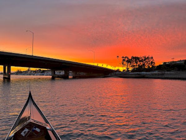 A boat on water approaches a bridge during a vibrant sunset, with colorful skies reflected on the calm surface, creating a serene scene.