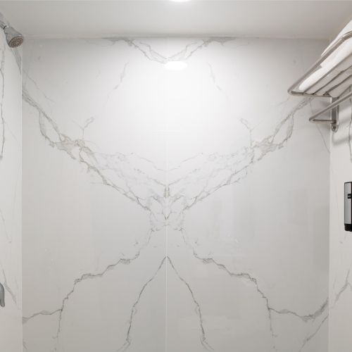 A marble-tiled shower with folded towels on a rack, a showerhead, a dispenser, and a curtain rod visible in the image.