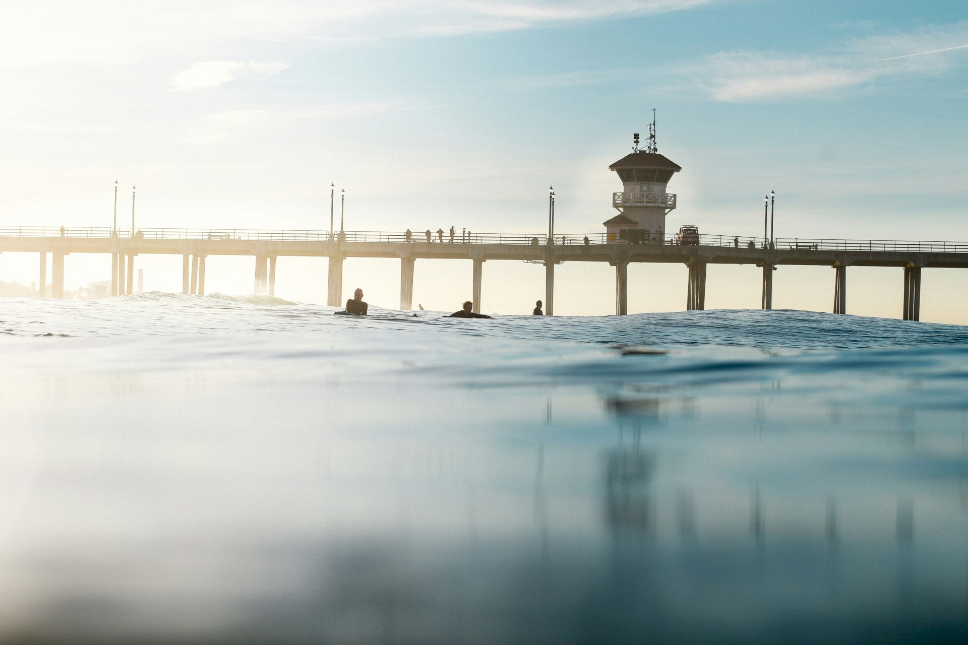 A serene image of surfers in the water with a pier and a tower in the background, under a clear sky.