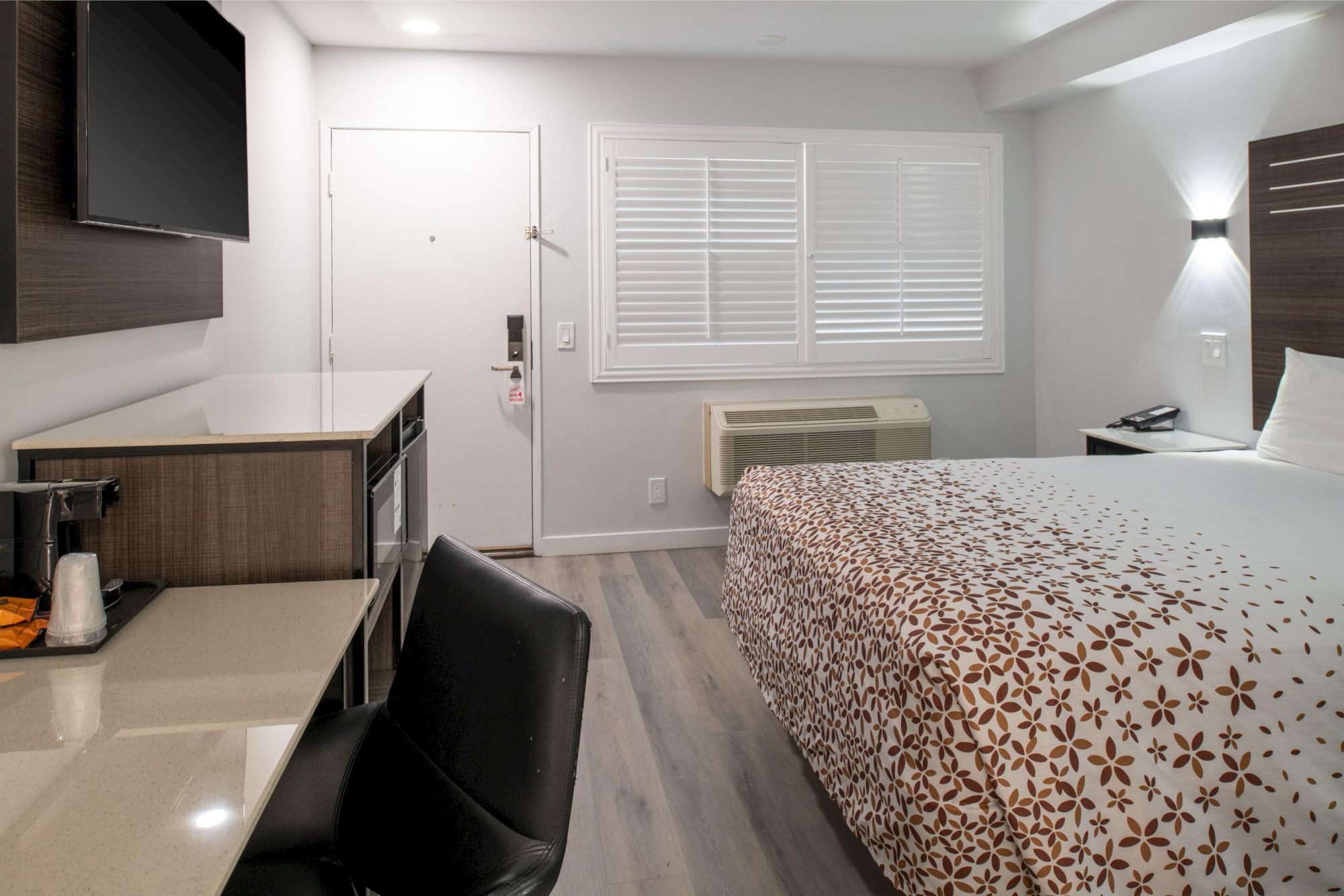 A tidy hotel room with a bed, desk, TV, and window. Decor is modern with neutral tones and simple patterns.