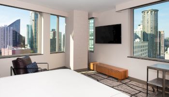 A modern hotel room with large windows showcasing a city view, a wall-mounted TV, contemporary furniture, and a bed.