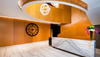 The image shows a modern lobby with a large wall clock, a marble reception desk, and wooden ceiling with decorative hanging lights.