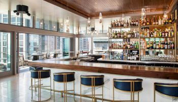 This image shows a modern bar with high chairs, hanging lights, and a wide selection of bottles, set in a room with large windows and city views.