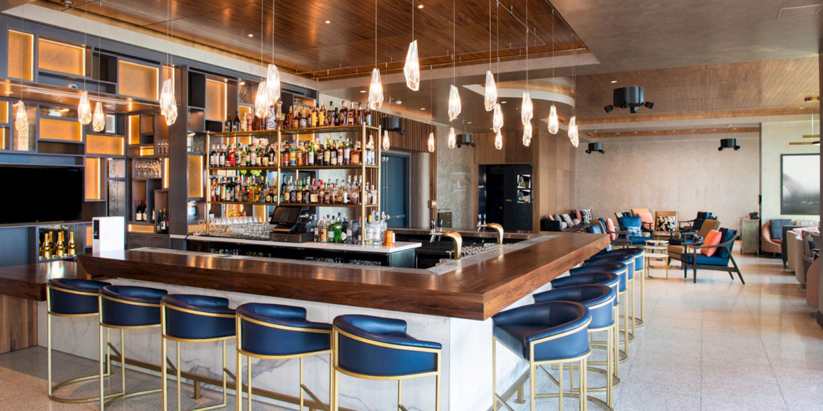 The image shows a modern bar with a wooden counter, blue stools, and a well-stocked bar. Hanging lights add ambiance to the spacious room.