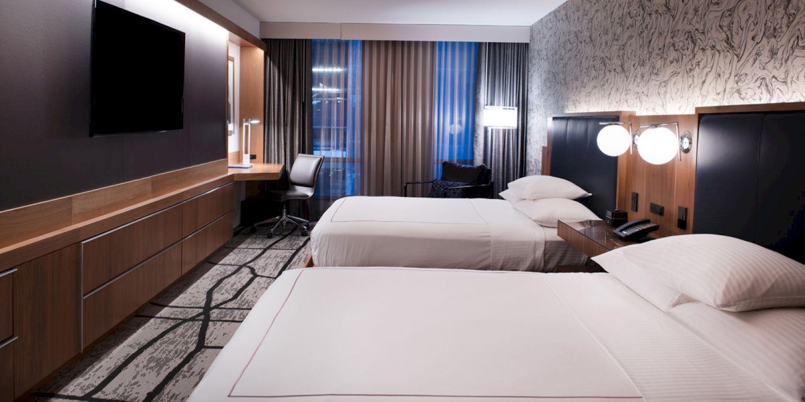A modern hotel room with two beds, a desk, a TV, and stylish lighting and decor finishes the look.