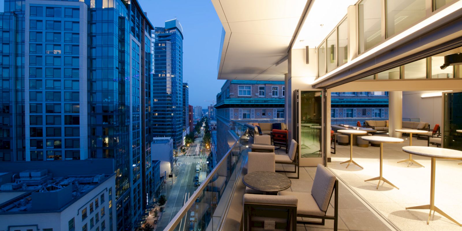 A modern balcony overlooks a cityscape with tall buildings at dusk, featuring outdoor seating and tables.