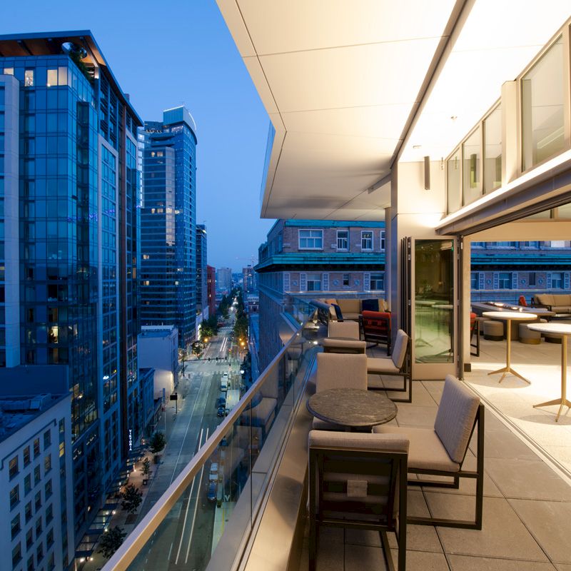 A rooftop terrace with seating overlooks a city skyline at dusk, with modern buildings and illuminated windows visible.