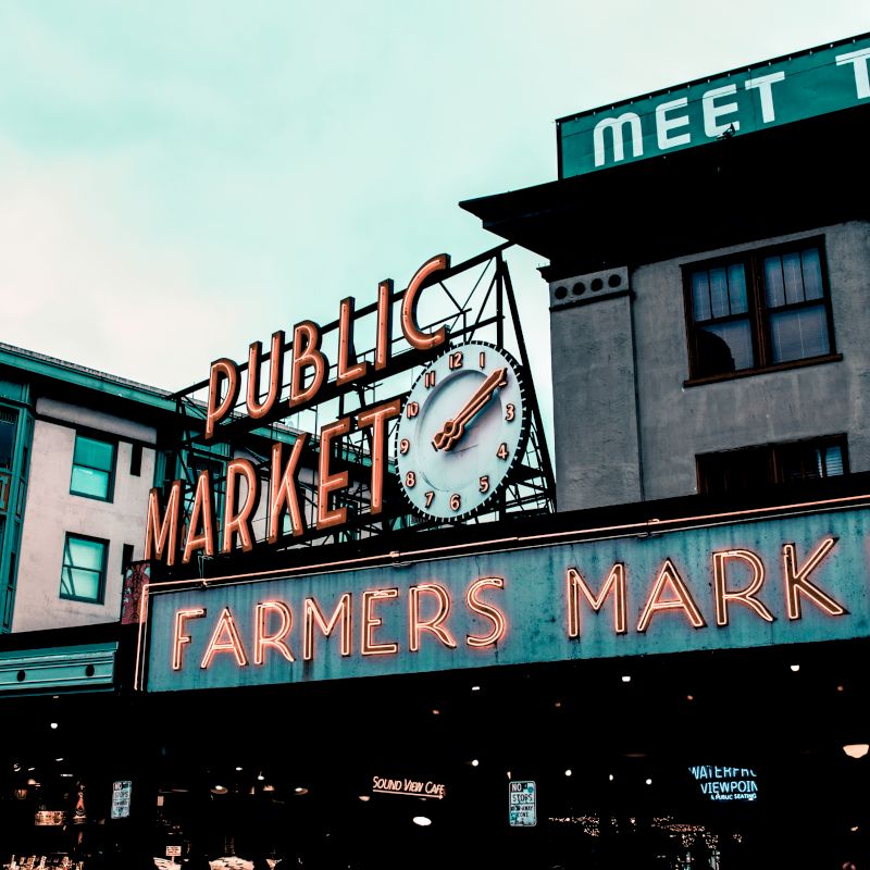 The image shows a neon sign with "Public Market" and "Farmers Market" displayed, along with a clock on a historic building.
