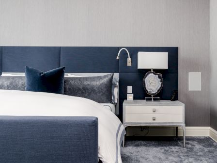 A modern bedroom with a blue and white color scheme. Features a bed with blue headboard, pillows, and a nightstand with a lamp and decor.