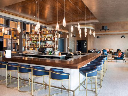 Modern bar with a central countertop, blue chairs, pendant lights, and well-stocked shelves. Cozy seating area in the background.